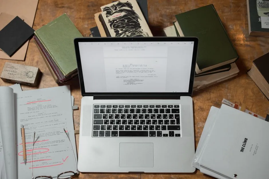 A laptop surrounded by books and scripts on a wooden desk, perfect for literature and technology themes.